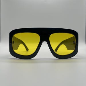 May include: Black oversized sunglasses with yellow lenses. The sunglasses have a thick, rectangular frame and a bold, modern design. The lenses are a bright, vibrant yellow, offering a striking contrast to the black frame. A stylish accessory for sun protection.