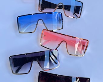 May include: Six pairs of oversized square sunglasses with gold frames and different colored lenses. The lenses are gradient, with a darker shade at the top and a lighter shade at the bottom. The sunglasses are arranged in a stack, with the darkest lenses at the bottom and the lightest lenses at the top.