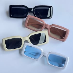 May include: Four pairs of sunglasses with different colored frames and lenses. The sunglasses have a square shape and are arranged in a row. The frames are black, pink, cream, and white. The lenses are black, brown, cream, and blue.
