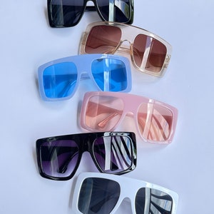 May include: A set of six sunglasses with different colored frames and lenses. The frames are all square shaped and the lenses are all dark. The sunglasses are arranged in a stack, with the black frames on top and the white frames on the bottom.