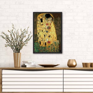 Op de afbeelding: Een ingelijste print van een schilderij dat twee figuren in een hartstochtelijke omhelzing toont. De figuren zijn gekleed in gouden en groene gewaden, en de achtergrond is een mozaïek van kleurrijke vierkanten. Het schilderij is getiteld "De Kus" van Gustav Klimt.