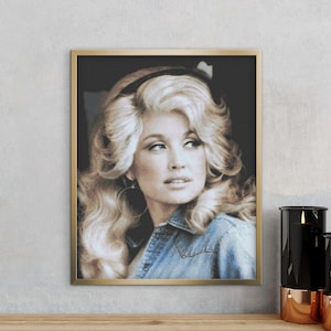 May include: A framed portrait print featuring a black and white stylized illustration of a person with voluminous blonde hair, wearing a denim shirt. The artwork is encased in a thin gold-toned frame, capturing a detailed facial expression.