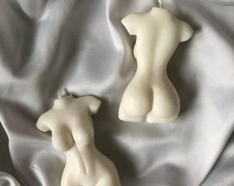 May include: Two white candle sculptures in the shape of a human torso. The candles are on a white satin fabric background.