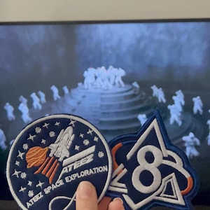 Puede incluir: Dos parches bordados con temática espacial. Uno es un círculo azul marino con un cohete y el texto "ATEEZ SPACE EXPLORATION". El otro es un parche azul y blanco con el número "8".