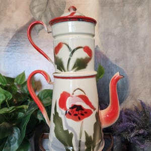 May include: A vintage white and red enamelware coffee pot with a floral design. The pot features a red handle, spout, and rim, with a lid. The floral design includes red poppies and green leaves. The coffee pot is displayed on a metal stand.