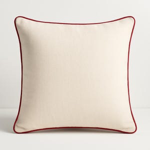 May include: Square cream cushion with a dark red trim. The cushion has a plain cream-coloured centre and a thin, dark red border around the edges. This cushion is likely for home decor and measures approximately 40cm x 40cm.