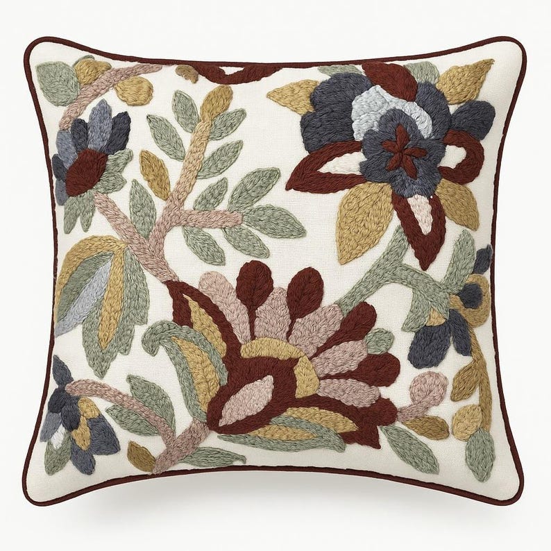 May include: Square cushion with an embroidered floral design. The cushion has a white background with a detailed pattern of flowers and leaves in shades of grey, brown, yellow, and green. It has a dark brown border.