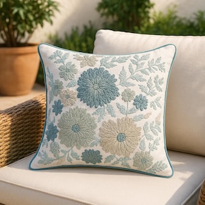 Op de afbeelding: Vierkant decoratief kussen met geborduurd bloemmotief in blauw- en beigetinten op een witte achtergrond. Het kussen heeft een turquoise rand en ligt op een neutraal gekleurd kussen.