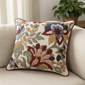 May include: Square cushion with an embroidered floral pattern. The cream-coloured fabric is adorned with colourful flowers in shades of red, blue, yellow, and green. It has a dark red border around the edges.
