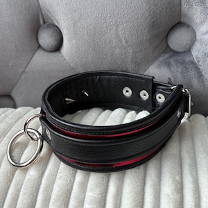 May include: A black leather collar with a red interior and silver-tone metal hardware. The collar has a large metal ring and studded details. It is displayed on a white, textured surface, likely a cushion.