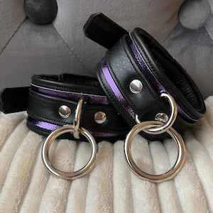 May include: Two black leather wrist cuffs with silver metal rings and a purple stripe. The cuffs are on a white fluffy surface.