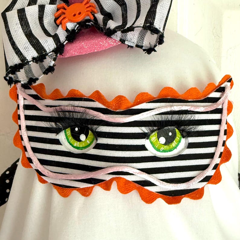 May include: A Halloween decoration featuring a sleep mask with black and white stripes, large green eyes, and long black eyelashes. The mask has orange scalloped edges and a pink string. A black and white striped bow with an orange spider is at the top.