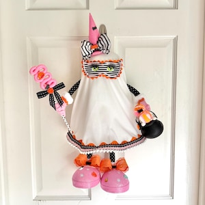May include: A Halloween ghost decoration with a pink witch hat, striped bow, and a mask with green eyes. It holds a pink lollipop and a black cauldron. The ghost is dressed in a white sheet with orange and black trim.
