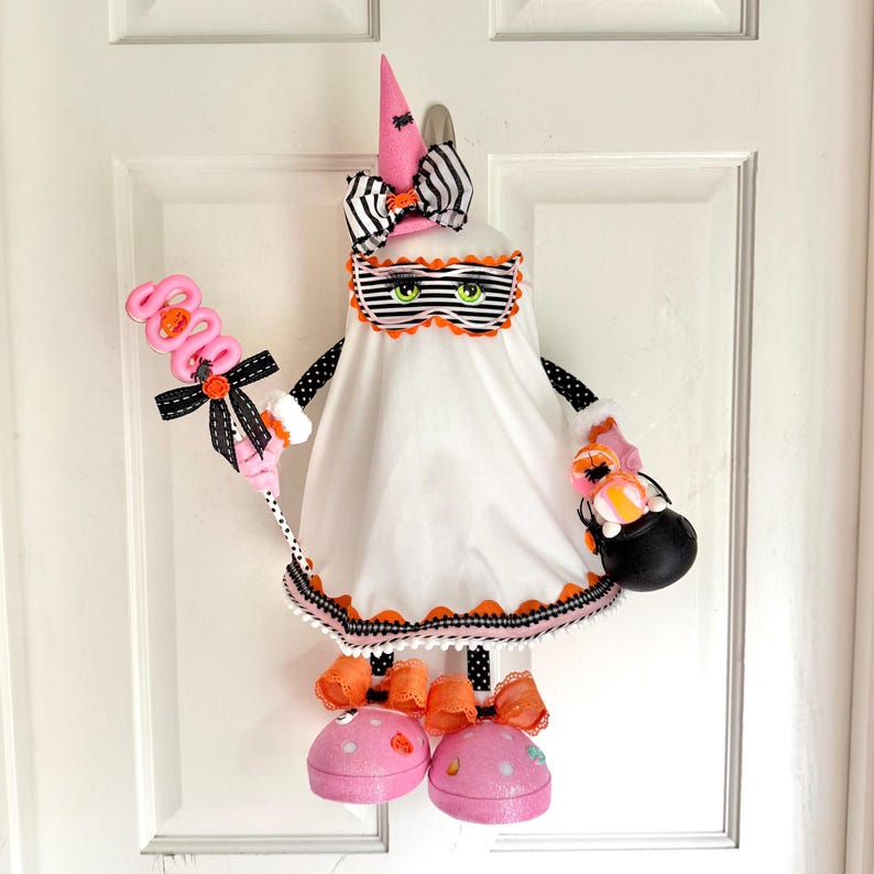 May include: A Halloween ghost decoration with a pink witch hat, striped bow, and a mask with green eyes. It holds a pink lollipop and a black cauldron. The ghost is dressed in a white sheet with orange and black trim.