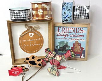 May include: A selection of dog-themed decorations. The image shows spools of ribbon with paw prints and bone designs, framed signs with dog-related quotes, and decorative paw print and dog bone picks. One sign reads "Friends always welcome".