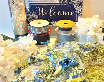 May include: A selection of craft supplies, including rolls of deco mesh, decorative ribbons, and a "Welcome" sign. The sign is navy blue with white lettering and floral accents. Artificial white and blue flowers and greenery are also visible.