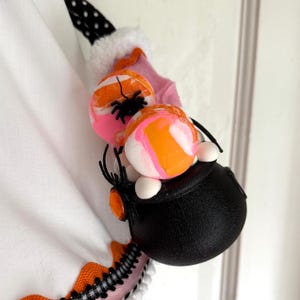 May include: A Halloween decoration with a black cauldron filled with orange, pink, and white candy-style spheres, decorated with black plastic spiders. The design incorporates pink fabric and a black and white polka dot detail.