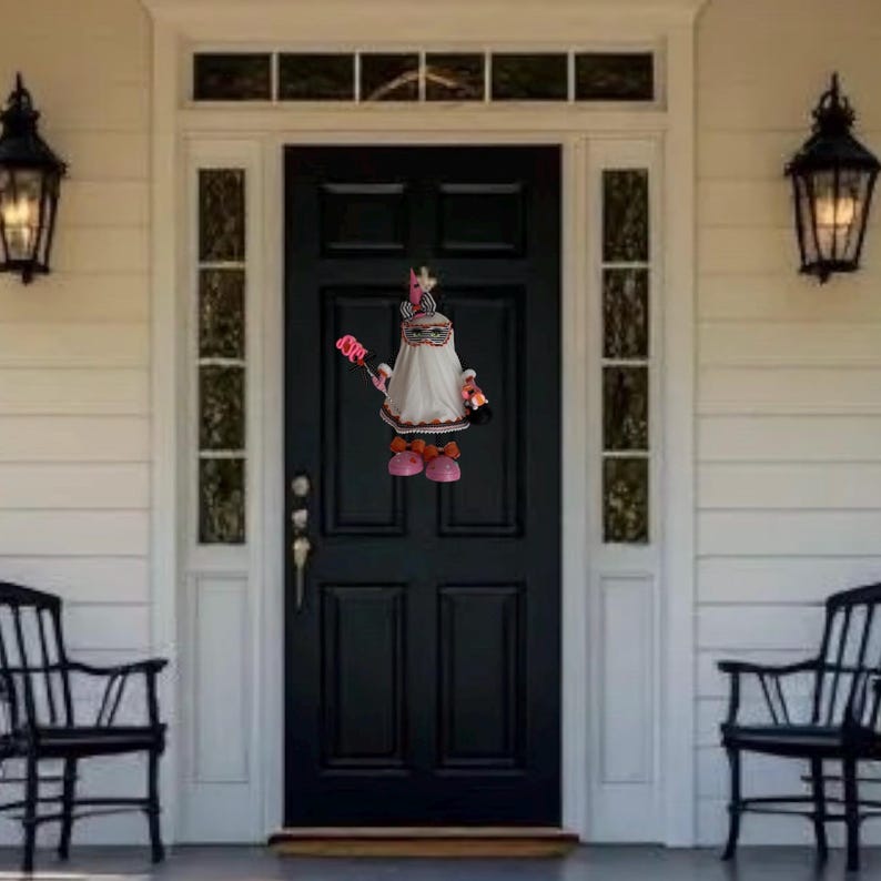 May include: A decorative doll with a white dress, pink shoes, and a headpiece, standing in front of a black door. The doll is holding a red and pink object. The door is framed by white trim and black lanterns. The scene is set outside.
