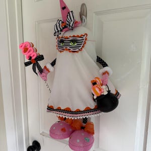 May include: A Halloween ghost decoration with a pink witch's hat and a striped mask. The ghost holds a pink lollipop and a black cauldron filled with treats. It has pink polka-dot shoes and is decorated with black and orange accents. The ghost is white.