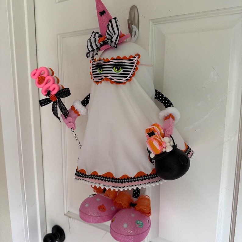 May include: A Halloween ghost decoration with a pink witch's hat and a striped mask. The ghost holds a pink lollipop and a black cauldron filled with treats. It has pink polka-dot shoes and is decorated with black and orange accents. The ghost is white.