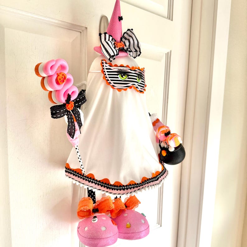 May include: A Halloween ghost decoration featuring a pink witch's hat, a striped bow, and a decorative mask with green eyes. The ghost wears a white dress with orange and black trim, holding a candy-filled treat bag and a pink wand.