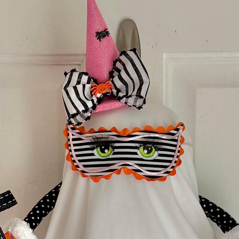 May include: A Halloween ghost decoration featuring a pink glitter witch's hat, a black and white striped bow, and a striped eye mask with green eyes. The ghost has black polka dot arms.