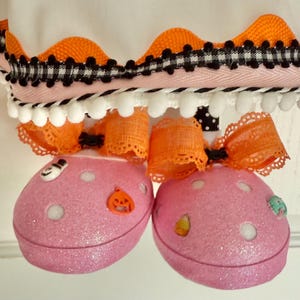 May include: A close-up of a Halloween decoration featuring two pink, glittery, rounded ornaments with white dots and small decorative buttons. The ornaments are adorned with orange lace bows and are connected to a decorative trim with orange and black accents.