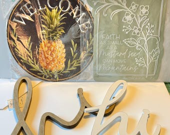 May include: A selection of decorative signs. A round sign with a pineapple design and the word "WELCOME". A rectangular sign with floral accents and the text "Faith as small as a mustard seed can move mountains". Wooden "hi" and "lo" signs are also present.
