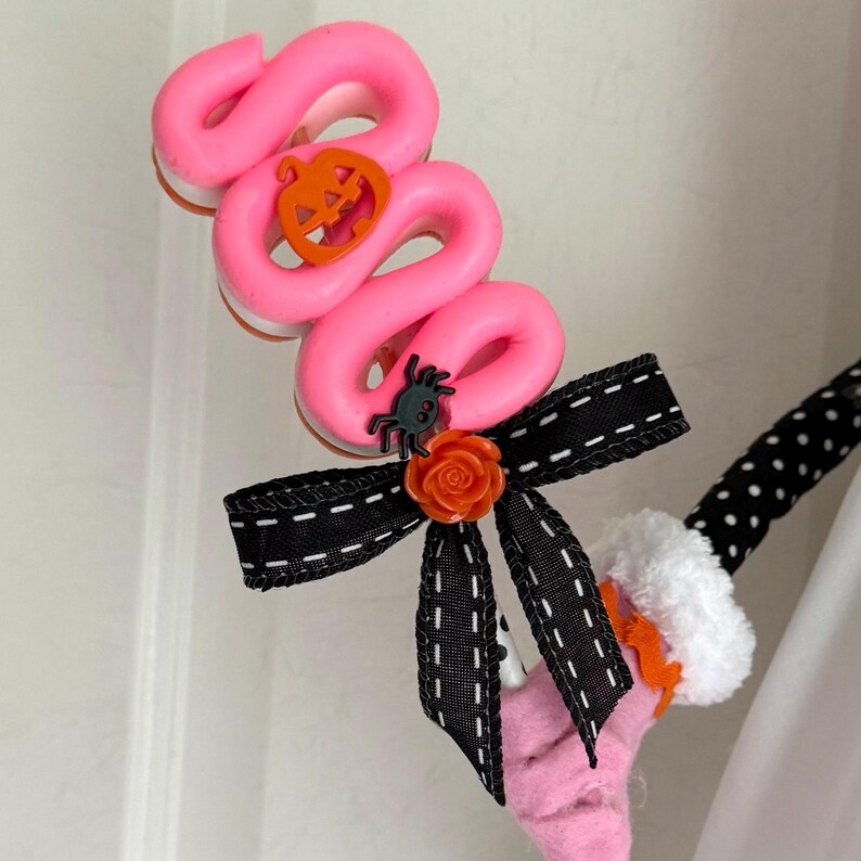 May include: A pink Halloween hair accessory with a swirled design, an orange pumpkin, and a black bow with white stitching. A small spider and orange rose add detail. The accessory has a black and white polka dot stem.