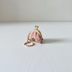 Op de afbeelding: Een kleine, roze munttasje-sleutelhanger met een goudkleurig metalen frame en ketting. De tas heeft een gouden bloemenpatroon en een sluiting met kliksluiting. De sleutelring is ook goudkleurig.
