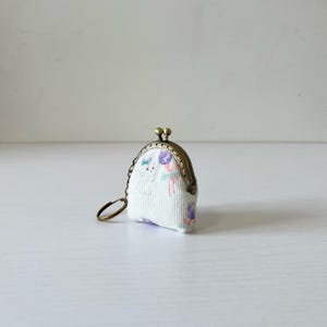 Puede incluir: Un pequeño monedero blanco con cierre metálico y llavero. El monedero presenta un estampado floral con detalles en morado, rosa y azul. El cierre y el llavero son de color bronce.