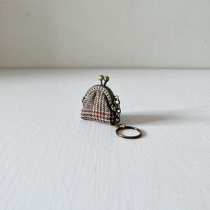 Op de afbeelding: Een kleine sleutelhanger met een muntportemonnee in vintage stijl. De portemonnee heeft een patroonstof in bruin-, rood- en wittinten, met een metalen sluiting en ketting. De sleutelring is ook van metaal. De portemonnee is een miniatuurversie van een klassiek ontwerp.