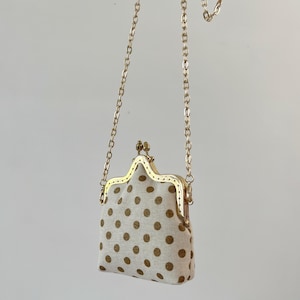 May include: A small, cream-colored purse with a gold chain strap. The purse features a gold clasp and is decorated with brown polka dots. The bag is suspended in the air against a plain background.
