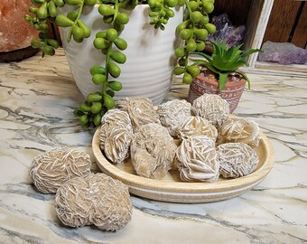 May include: A collection of tan-colored rose quartz crystals, also known as desert roses, arranged in a wooden bowl. The crystals have a unique, layered, and delicate appearance.