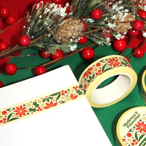 May include: Two rolls of washi tape with a Christmas theme. The tape features a repeating pattern of red poinsettias, green holly leaves, gold stars, and red berries on a cream background. The tape is rolled up and the ends are visible. The tape is on a green surface with red berries and a pine branch.