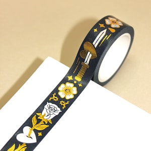May include: Black washi tape with gold foil accents. The tape features a repeating pattern of flowers, hearts, and a sword.