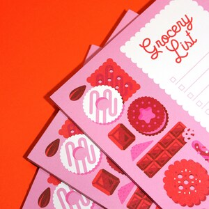 May include: Three pink grocery list notepads with a white border and a scalloped edge. The top notepad has the text "Grocery List" in red. The notepads are decorated with illustrations of cookies, chocolate bars, and other sweets.