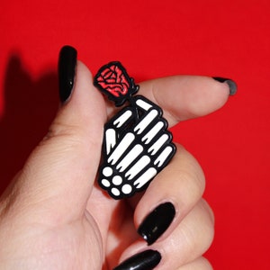 May include: A black enamel pin featuring a skeleton hand holding a red rose. The pin is on a person's hand with black nail polish.