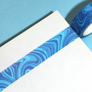 May include: A roll of blue decorative tape with a swirling pattern of light and dark blue lines. The tape is partially unrolled, lying diagonally across a white surface, against a light blue background. The tape is for crafts and decoration.