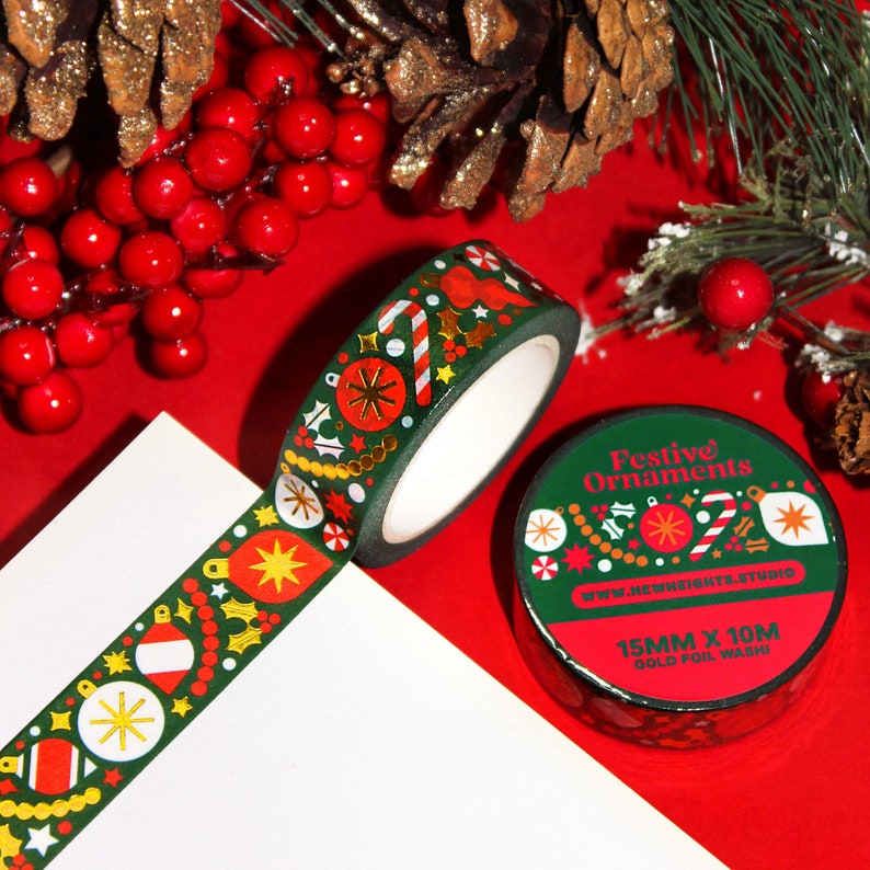 This Christmas washi tape features abstract holiday shapes of various styles of ornaments, garlands, tree decor, and mistletoes. This is a dark green washi tape, with white and red elements, and gold foiling details. Pictured is a roll and packaging.