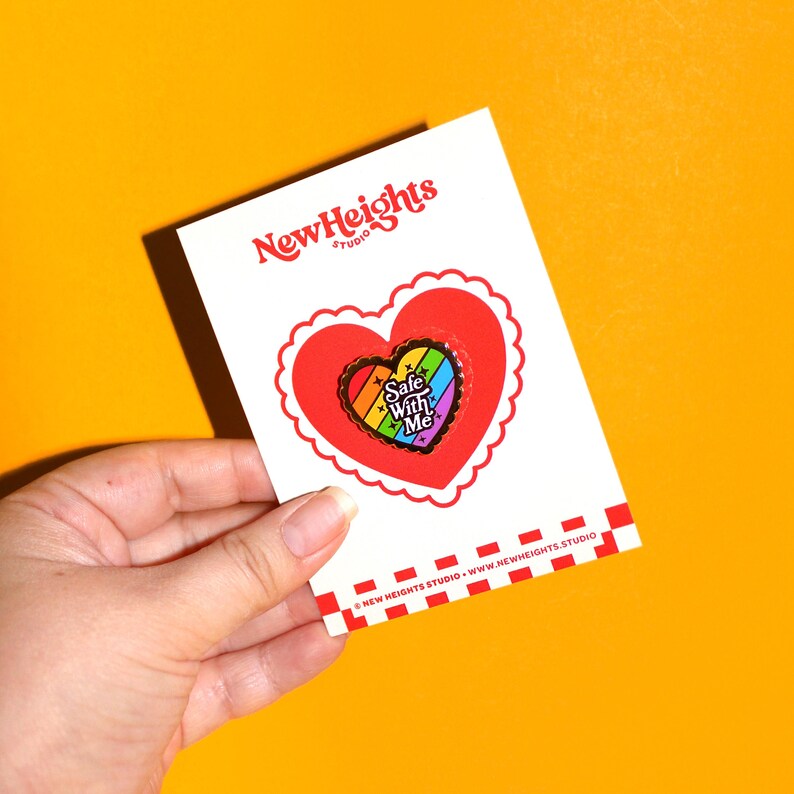 Op de afbeelding: Een witte kaart met een rode en zwarte geruite rand. De kaart heeft de tekst "New Heights Studio" in rood en zwart. Een rood hart met een witte gegolfde rand omringt een regenboog hart emaille pin met de tekst "Safe With Me".