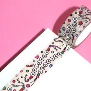 May include: A roll of beige washi tape with a colorful pattern of hearts, stars, smiley faces, and beads. The tape features the words "Friends Forever" and "I Love You" in white letters.