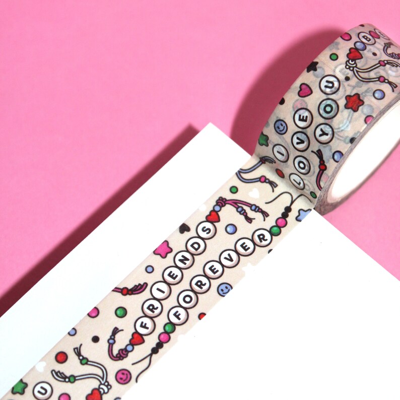 May include: A roll of beige washi tape with a colorful pattern of hearts, stars, smiley faces, and beads. The tape features the words "Friends Forever" and "I Love You" in white letters.
