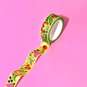 May include: A roll of green and yellow washi tape with a colorful fruit and vegetable pattern. The tape is unrolled on a pink background.