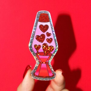 May include: A sticker of a lava lamp with a pink body and red glitter hearts. The base and top are red, and the lamp is outlined with a silver glitter border. The background is a solid red.
