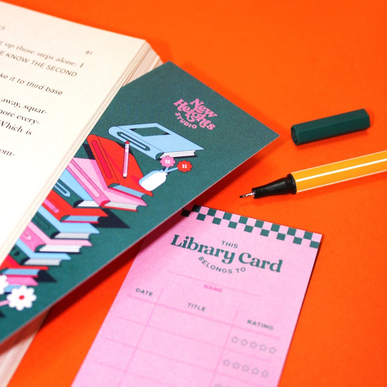 May include: A pink library card with the text "This Library Card Belongs To" and spaces for the borrower's name, date, title, and rating. The card has a checkered pattern in the top right corner. The card is on a bright orange surface.