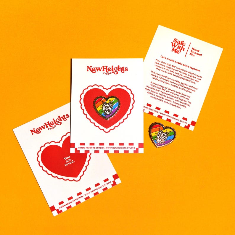 Op de afbeelding: Een regenboogkleurige, hartvormige emaille-speld met de tekst "Safe With Me" in witte letters. De speld bevindt zich op een witte kaart met de tekst "NewHeights Studio" en een kleinere regenboogkleurige, hartvormige speld met dezelfde tekst. De kaart bevat ook de tekst "Laten we samen een veiligere plek cre&euml;ren. Deze "Safe With Me" emaille-speld betekent simpelweg dat degenen die hulp, steun en bescherming zoeken, veilig bij jou zijn, ongeacht wie ze zijn of welke identiteiten ze hebben. Deze speld symboliseert de strijd tegen geweld tegen kwetsbare en gemarginaliseerde groepen en cre&euml;ert tegelijkertijd een veilige ruimte. Een deel van de opbrengst van onze "Safe With Me" speld gaat naar verschillende liefdadigheidsorganisaties die de strijd tegen racisme, seksueel geweld, geweld tegen vrouwen, homofobie, transfobie en saphophobie ondersteunen."