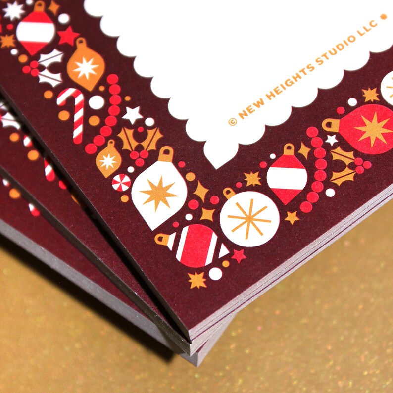 May include: A stack of Christmas cards with a burgundy border featuring festive ornaments, stars, and holly. The cards have a white scalloped edge and the text "NEW HEIGHTS STUDIO LLC" in gold.