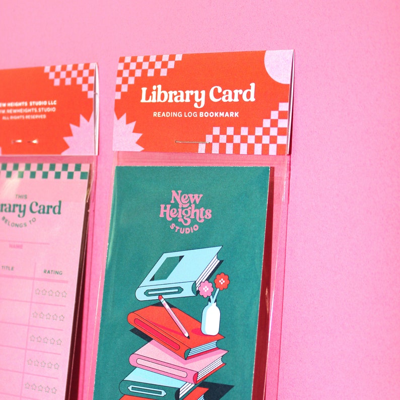 May include: A pink and green bookmark with the text "Library Card" and "Reading Log Bookmark" on the top. The bookmark features a stack of books with a pencil and a vase with flowers on top. The text "New Heights Studio" is on the bookmark.