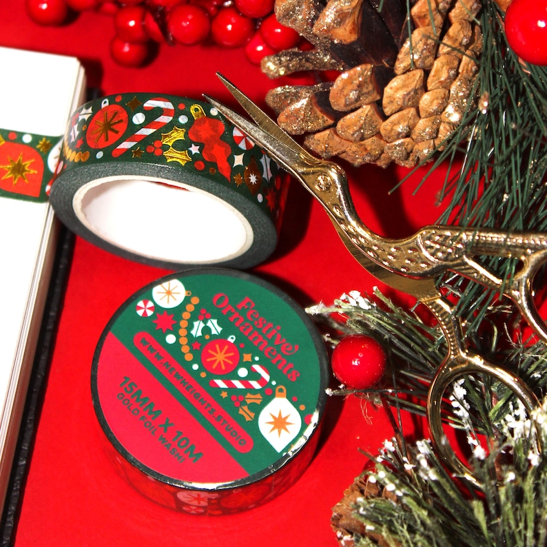 This Christmas washi tape features abstract holiday shapes of various styles of ornaments, garlands, tree decor, and mistletoes. This is a dark green washi tape, with white and red elements, and gold foiling details. Pictured is a roll and packaging.
